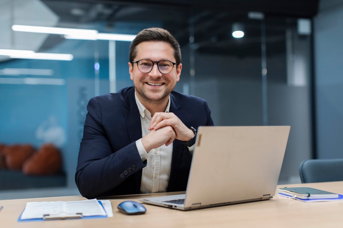 Portrait of successful businessman sitting at desk in office, working with documents and laptop, confidently smiling at camera