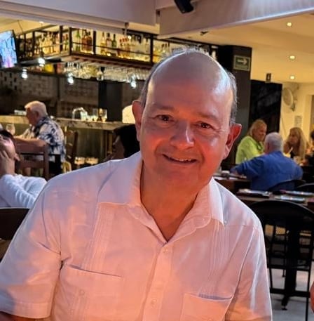 Middle-aged man smiling at camera in casual setting, wearing light pink button-up shirt in what appears to be a bar or restaurant